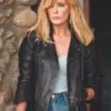 Beth Dutton wearing a black leather jacket and blue jeans standing against a stone wall.