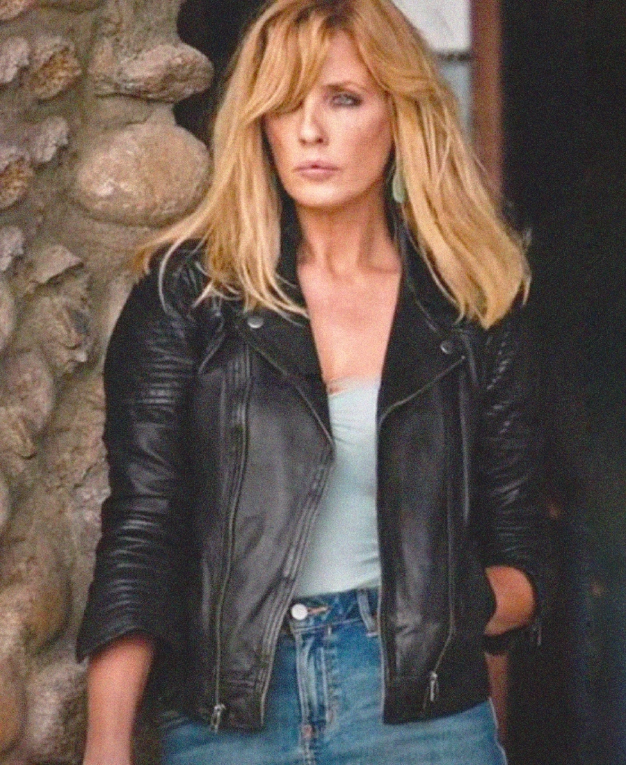 Beth Dutton wearing a black leather jacket and blue jeans standing against a stone wall.