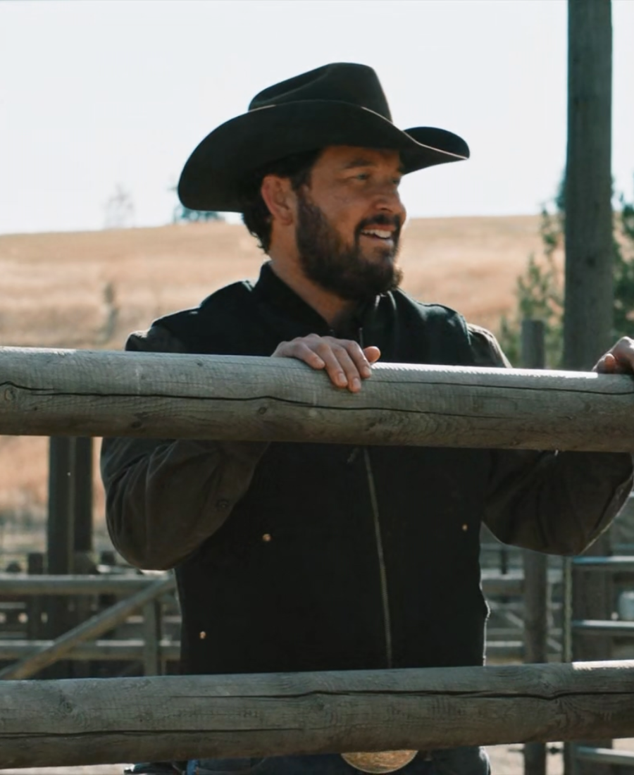 Rip Wheeler Wool Vest wearing a black cowboy hat and jacket standing behind a wooden fence.