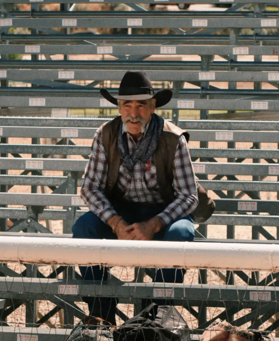 Yellowstone S04 Lloyd Vest Man in cowboy hat and plaid shirt sitting on metal cattle pens.
