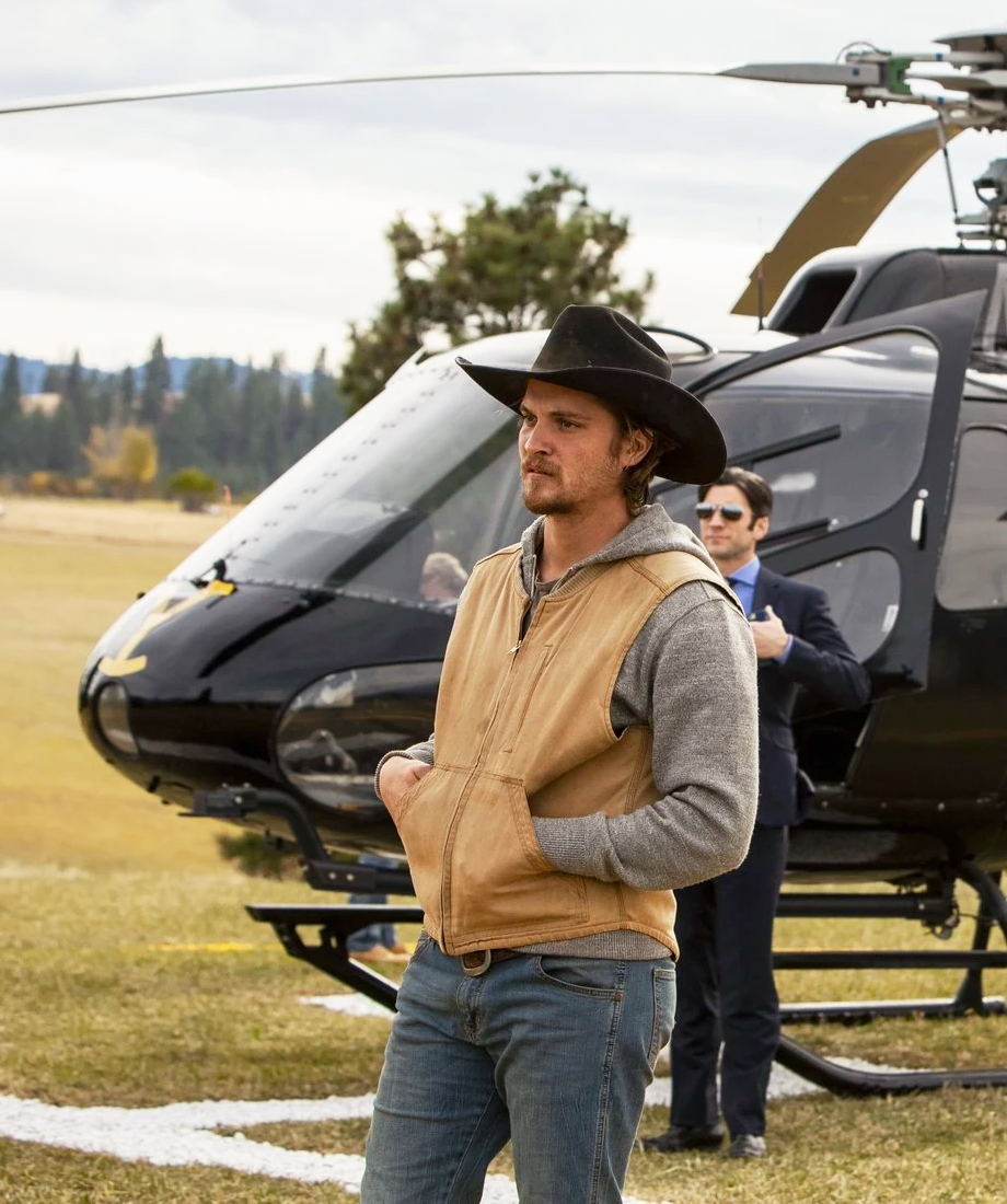 Man in a cowboy hat and vest standing in front of a helicopter on a grassy field.