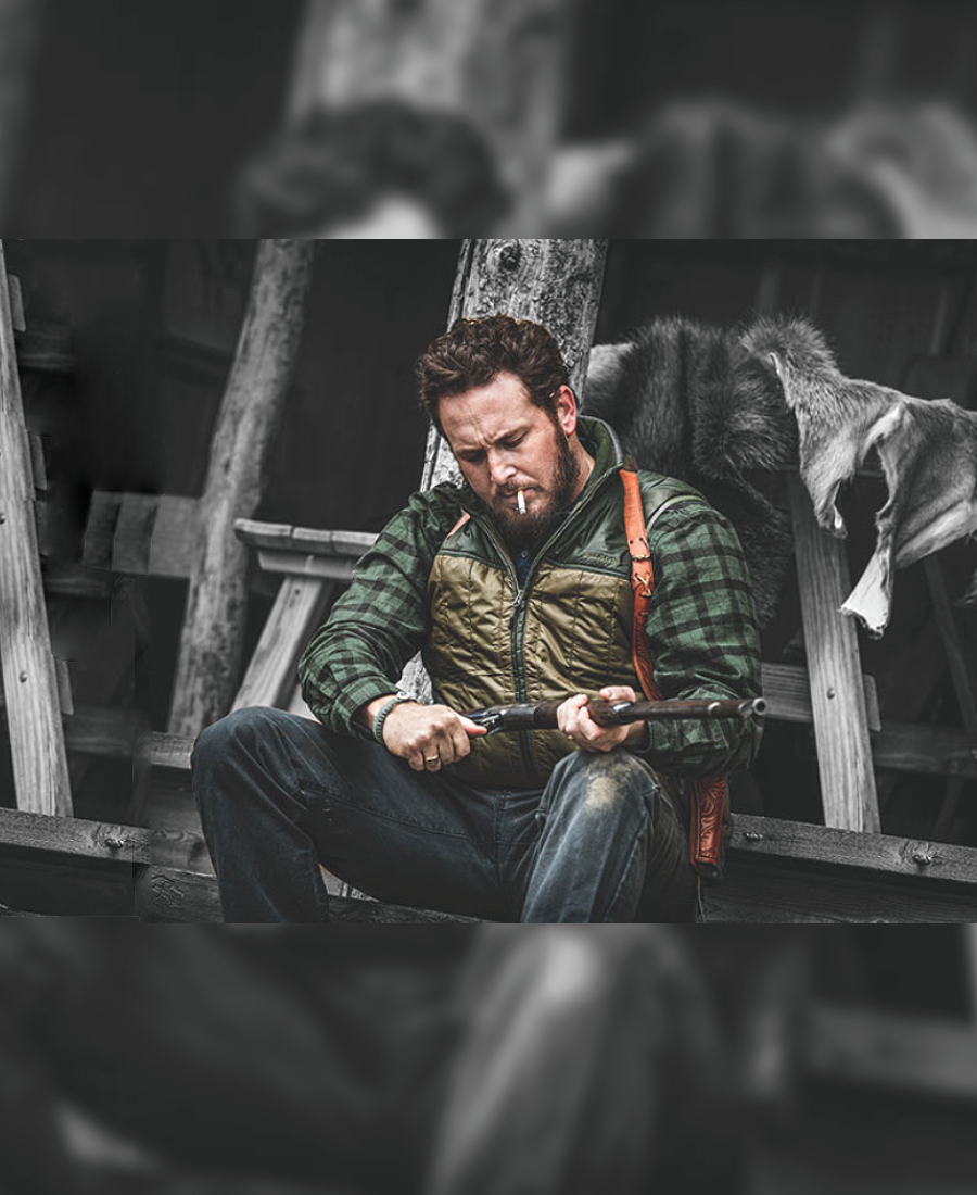 Rip Wheeler sitting on a wooden platform holding a tool, surrounded by rustic elements.