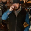 Rip Wheeler Black Wool vest with cowboy hat drinking from a glass in a rustic setting with wooden walls and horse equipment.