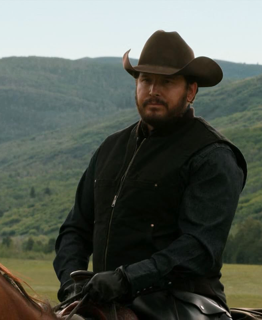 Rip Wheeler in a cowboy hat and black wool vest riding a horse with mountains in the background In Yellowstone Movie