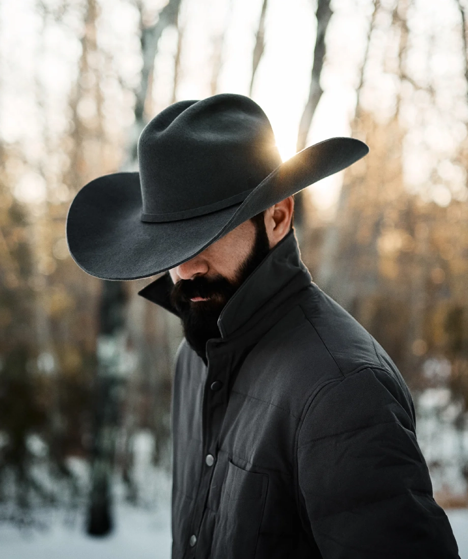 Man wearing a black cowboy hat and coat in a snowy forest